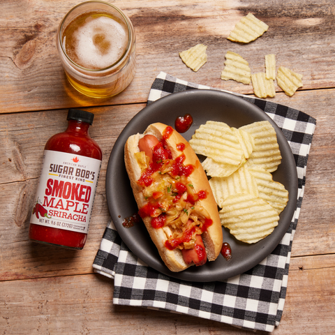 Hot dog with smoked maple sriracha sauce on a plate with chips and a drink on a wooden table.