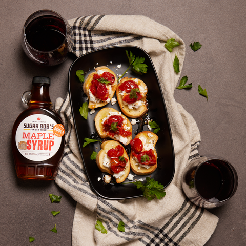 Maple syrup bottle, crostini with tomato and cheese on a platter, glasses of red wine, and checkered towel on a gray surface.