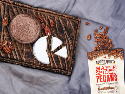 A wheel of baked Brie cheese topped with maple spiced pecans, served with crackers and apple slices on a wooden board.